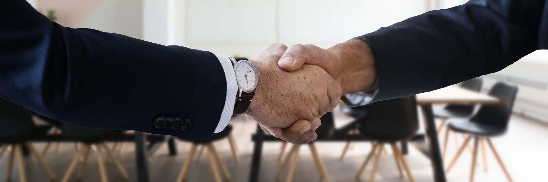 Händedruck Zwei Personen schütteln sich die Hand, dahinter ein Konferenztisch