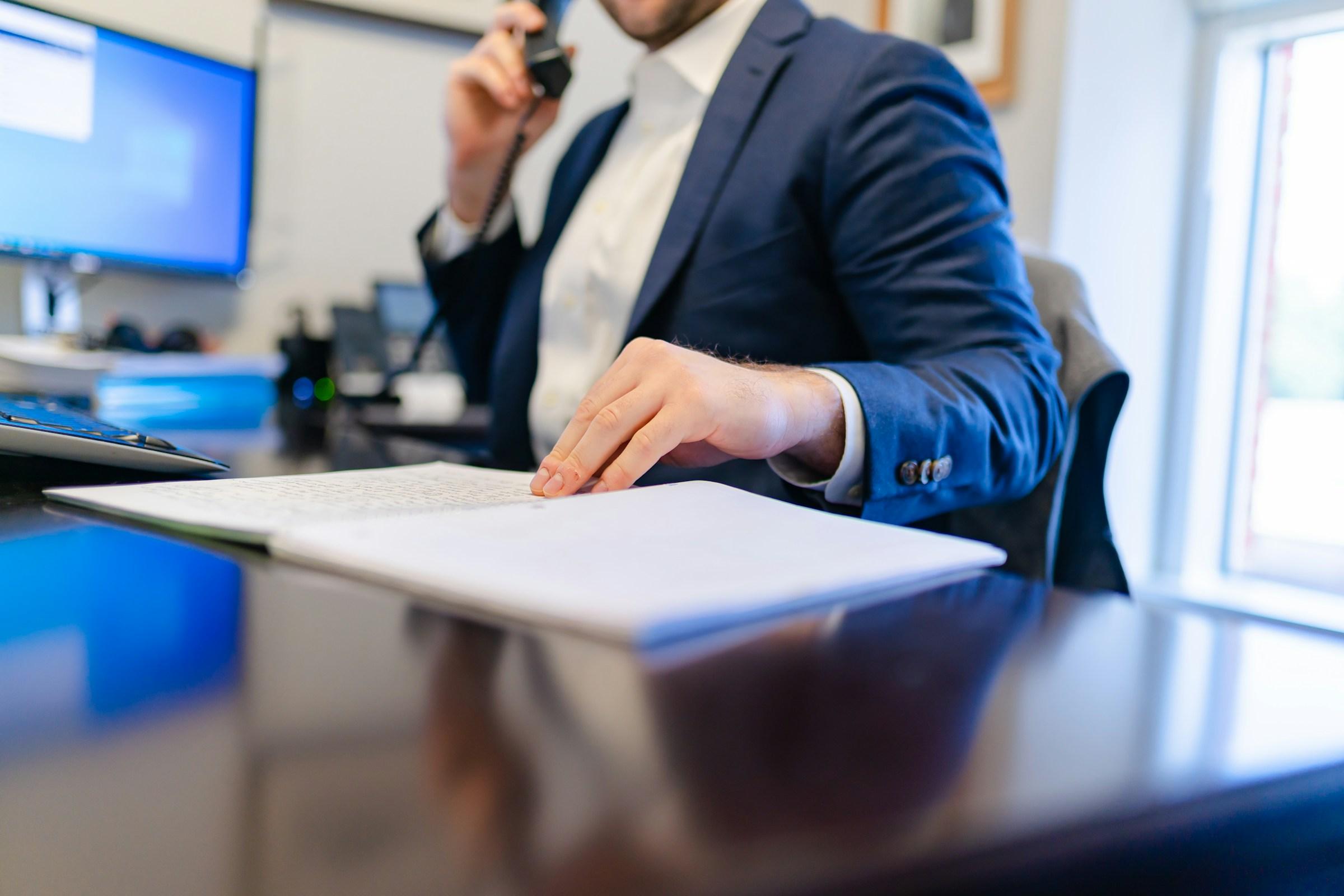 Gespräch Man in a suit sits in front of a computer with a telephone receiver in his hand and leafs through documents.